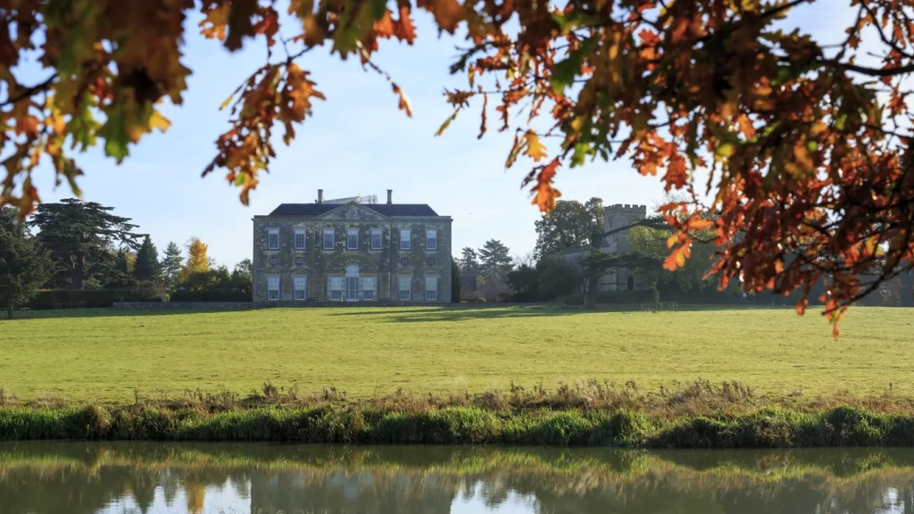 Georgian stone manor house viewed across a lake, framed by autumn oak leaves
